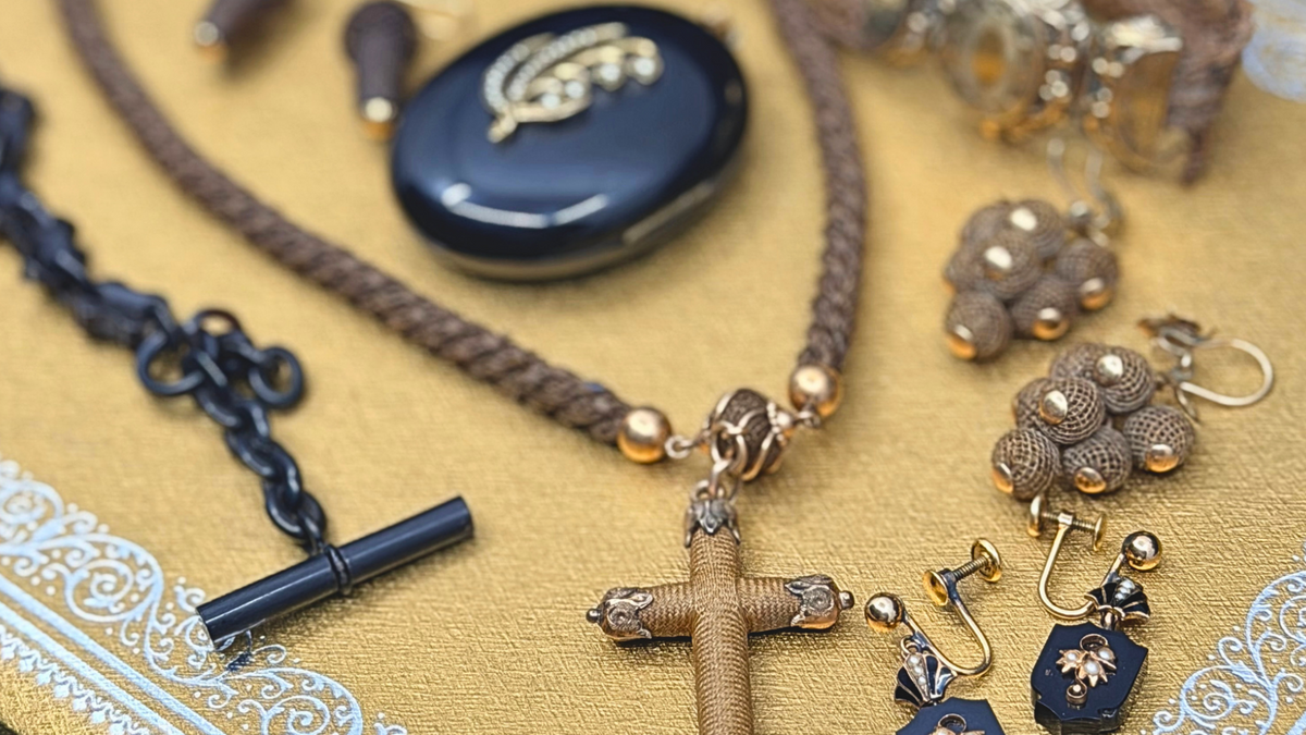 A styled flat-lay of Victorian mourning and hair jewelry on a mustard-yellow background, featuring a braided brown hair necklace, a jet watch chain, and a other woven hair earrings. 